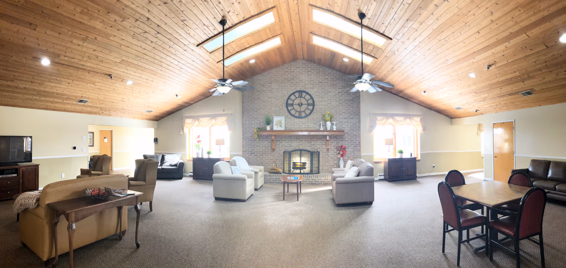 Spacious senior living common room with a vaulted wood ceiling, central brick fireplace, multiple seating areas and a dining table.