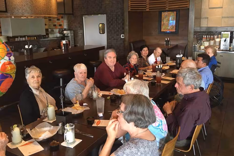 A group of elderly people sitting around a long dining table in a restaurant or dining area, enjoying drinks and food. The setting includes wooden furniture, a counter with stools, and a beverage station in the background.