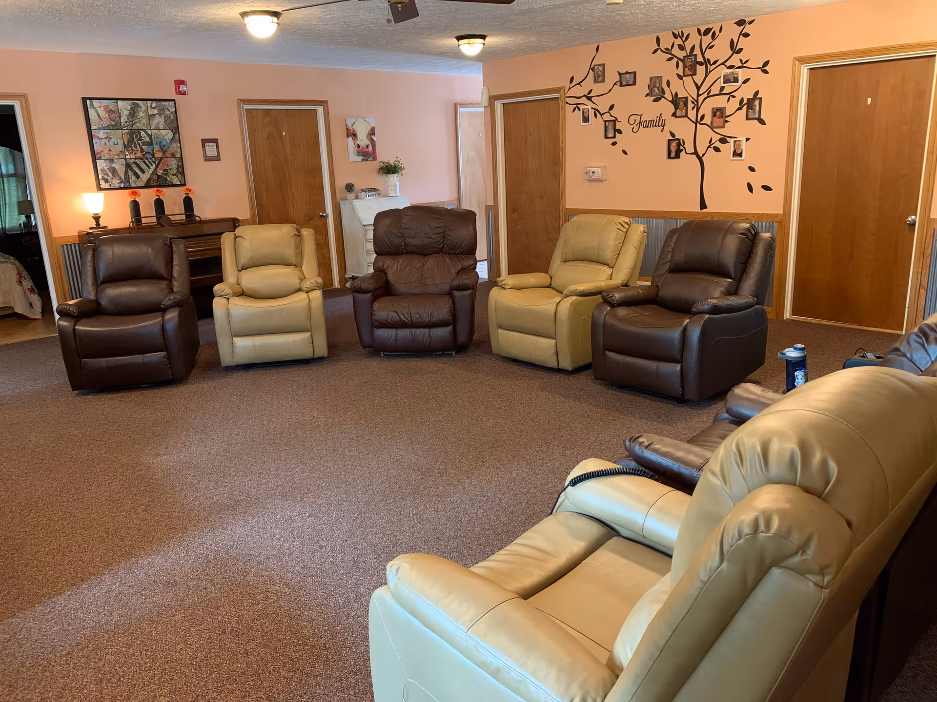 A communal living room with several beige and brown recliner chairs arranged in a semicircle and a 'Family' tree wall decal on a peach-colored wall.