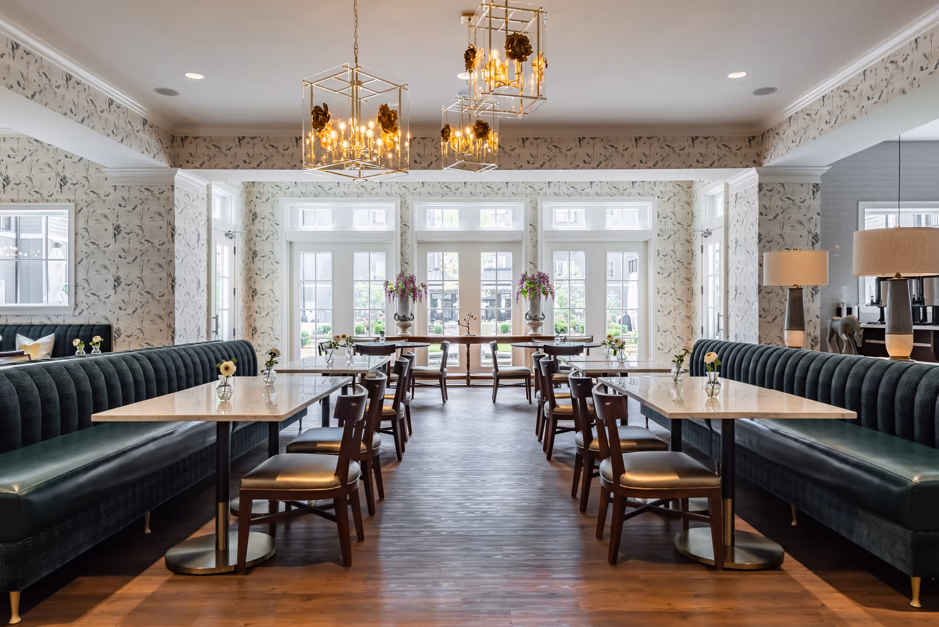 A bright and elegant dining room with marble-top tables, dark green upholstered bench seating along the walls, wooden chairs, and floral wallpaper. Large windows and glass doors at the far end let in natural light, and decorative chandeliers hang from the ceiling. Small vases with flowers are placed on each table.