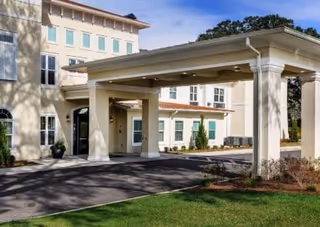 Exterior view of The Blake at Pensacola senior living facility showing the entrance with a covered driveway, beige building walls, multiple windows, and a well-maintained lawn.
