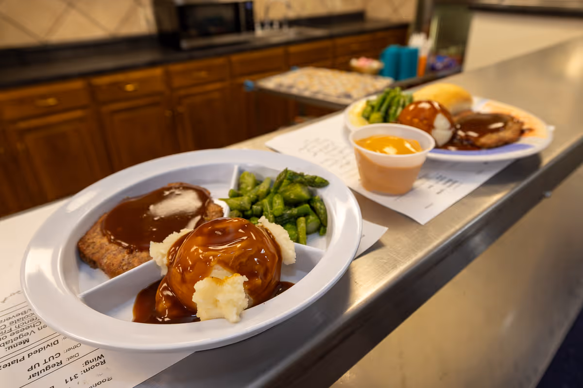 Two white divided plates on a metal counter in a kitchen setting, each plate containing a meal with mashed potatoes covered in brown gravy, green asparagus, a meat patty with brown gravy, and a bread roll. A small cup of orange sauce is also on one of the plates. Wooden cabinets and kitchen appliances are visible in the background.