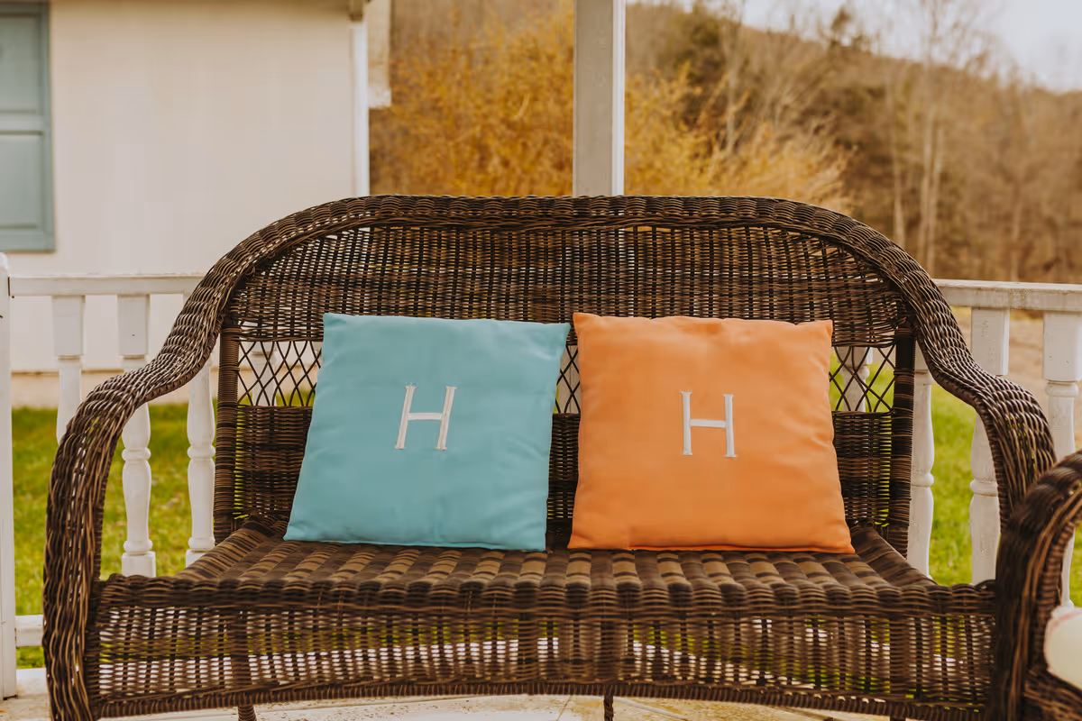 A brown wicker bench on a porch with two square cushions, one light blue and one orange, each embroidered with a white letter 'H'. The porch has white railings and a view of green grass and trees in the background.