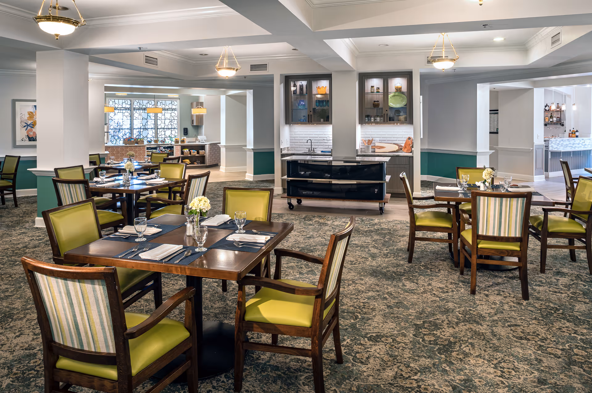 A spacious dining room with multiple wooden tables and chairs featuring green and striped upholstery. Each table is set with placemats, napkins, glassware, and a small floral centerpiece. The room has a patterned carpet, white walls with green wainscoting, and ceiling lights. In the background, there is a serving area with shelves holding dishes and glassware.