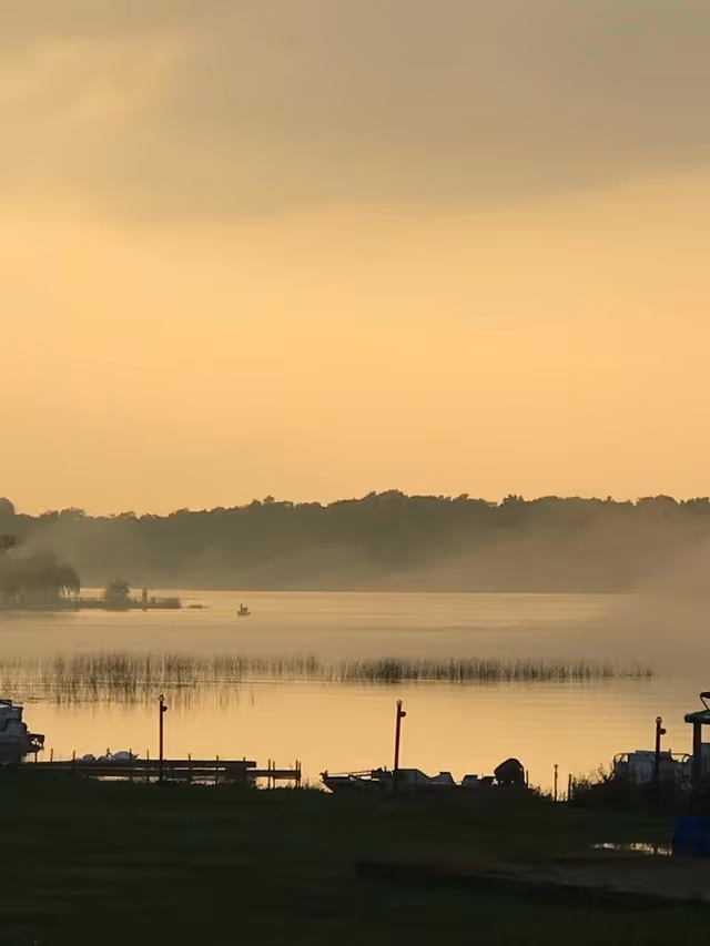 A calm waterfront scene at sunset or sunrise with a misty lake, silhouetted trees in the background, and small docks along the shore in the foreground.
