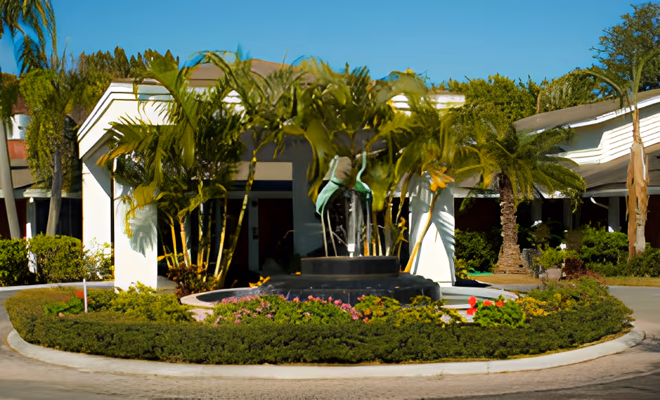 Entrance of a senior living facility with a circular driveway and a landscaped roundabout featuring a fountain surrounded by palm trees and flowering plants under a clear blue sky.