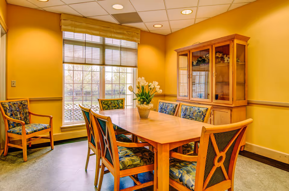 A dining room with a wooden table surrounded by six chairs with patterned cushions. A vase with white flowers is placed in the center of the table. There is a large window with blinds partially open, allowing natural light to enter the room. A wooden cabinet with glass doors is against the wall, displaying decorative items inside. The walls are painted yellow and the floor is a light gray color.