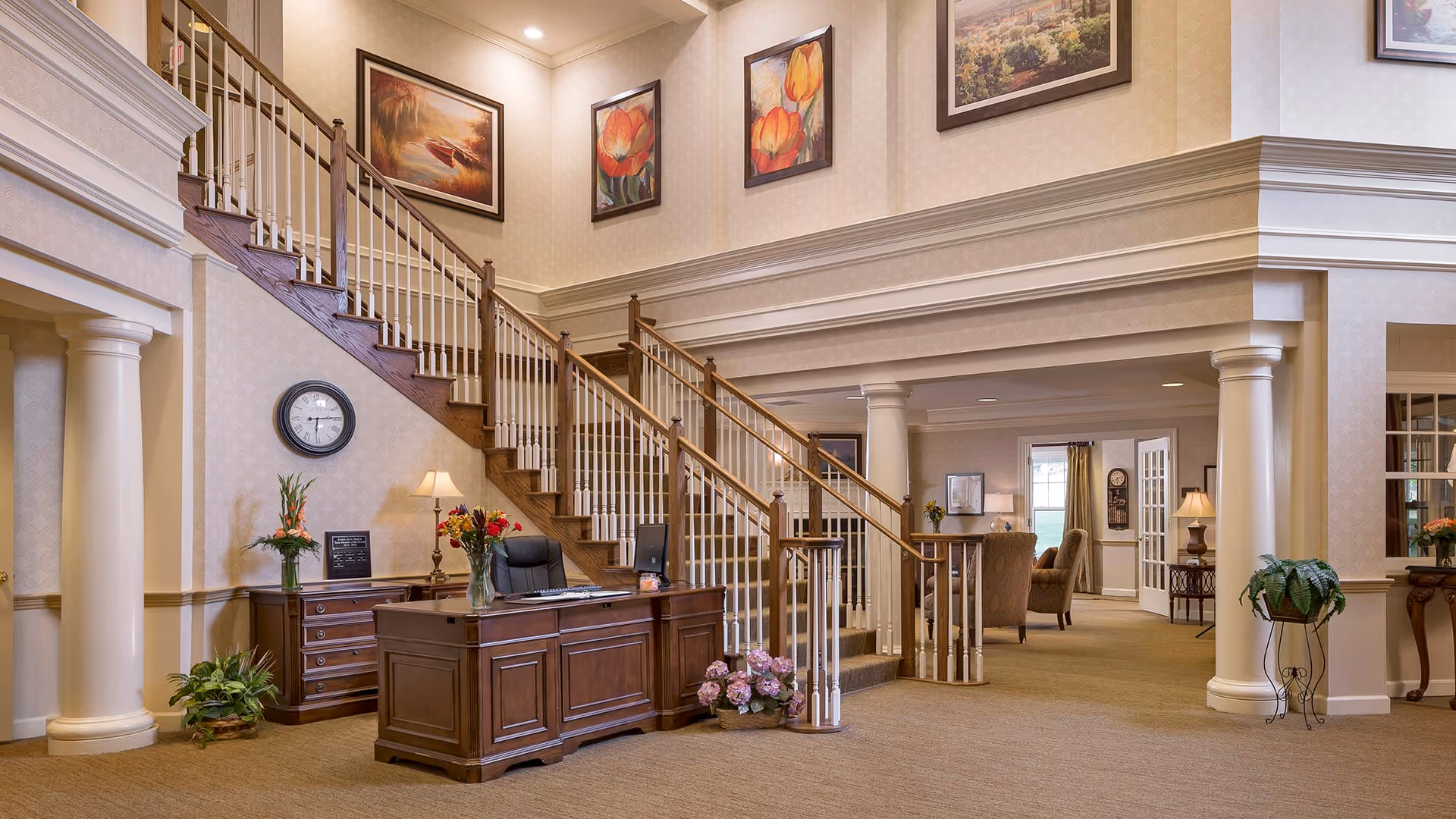 A spacious and elegant interior lobby area featuring a wooden reception desk with a computer and flower arrangements. Behind the desk is a staircase with wooden handrails and white balusters leading to an upper floor. The walls are decorated with framed artwork and a clock. The area is well-lit with lamps and recessed lighting, and there are columns and comfortable seating areas visible in the background.
