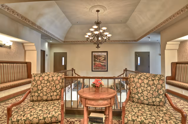 A cozy interior seating area with two floral upholstered armchairs and a small wooden round table with a flower arrangement in the center. Behind the seating area is a railing overlooking a lower level, with two doors and a framed picture on the wall. A decorative chandelier hangs from the ceiling.