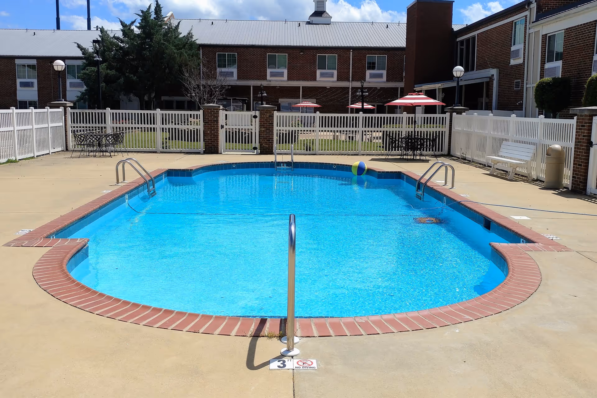 Outdoor swimming pool with clear blue water surrounded by a concrete deck and a white fence. There are metal handrails for entering the pool, a colorful beach ball floating in the water, and several tables with red and white umbrellas in the background. The pool area is part of a senior living community with brick buildings visible behind the fence.
