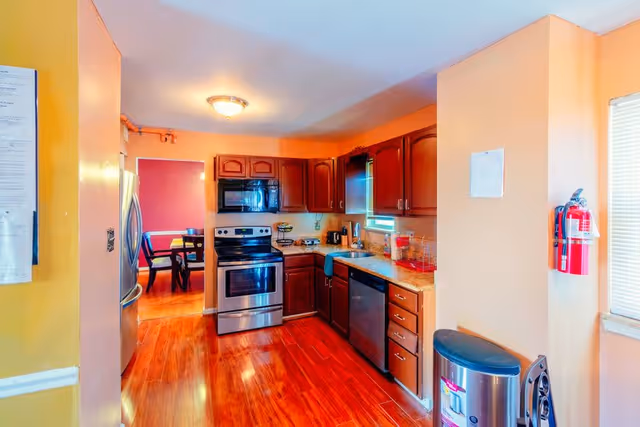 A bright kitchen with wooden cabinets, stainless steel appliances including a refrigerator, stove, and dishwasher. The kitchen has a granite countertop with a sink under a window with blinds. There is a fire extinguisher mounted on the wall and a trash can next to it. The floor is wooden and a dining area with a table and chairs is visible through an open doorway.