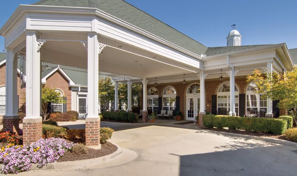 Entrance of a senior living facility with a covered driveway supported by white columns with brick bases. The building has a green roof, brick walls, and large arched windows with black shutters. There are neatly trimmed bushes and colorful flowers around the entrance area under a clear blue sky.