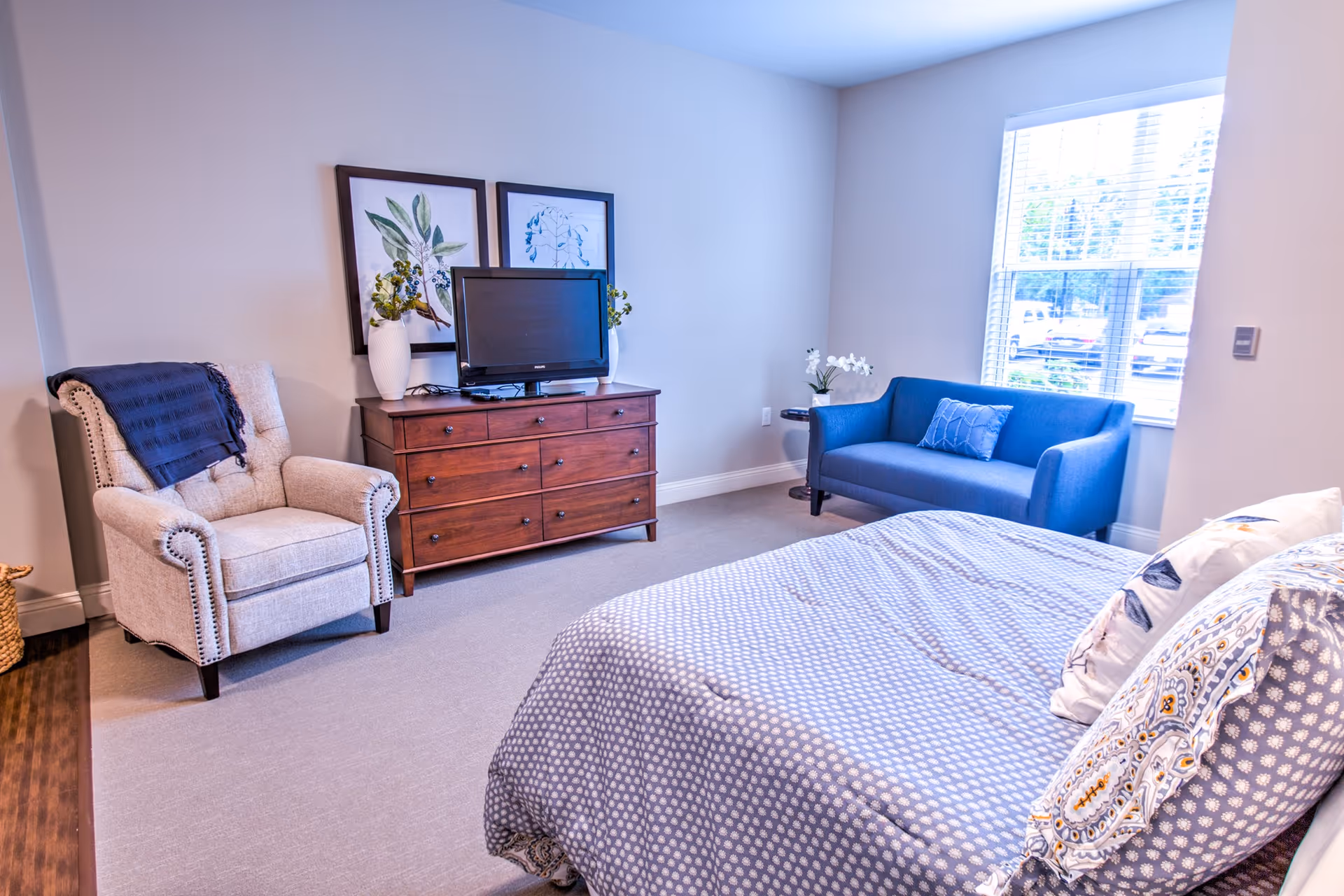 A cozy bedroom in a senior living facility featuring a bed with patterned bedding, a beige armchair with a dark throw blanket, a wooden dresser with a TV on top, two framed botanical prints on the wall, a blue loveseat with a decorative pillow, a small round side table with a white flower vase, and a window with blinds letting in natural light.