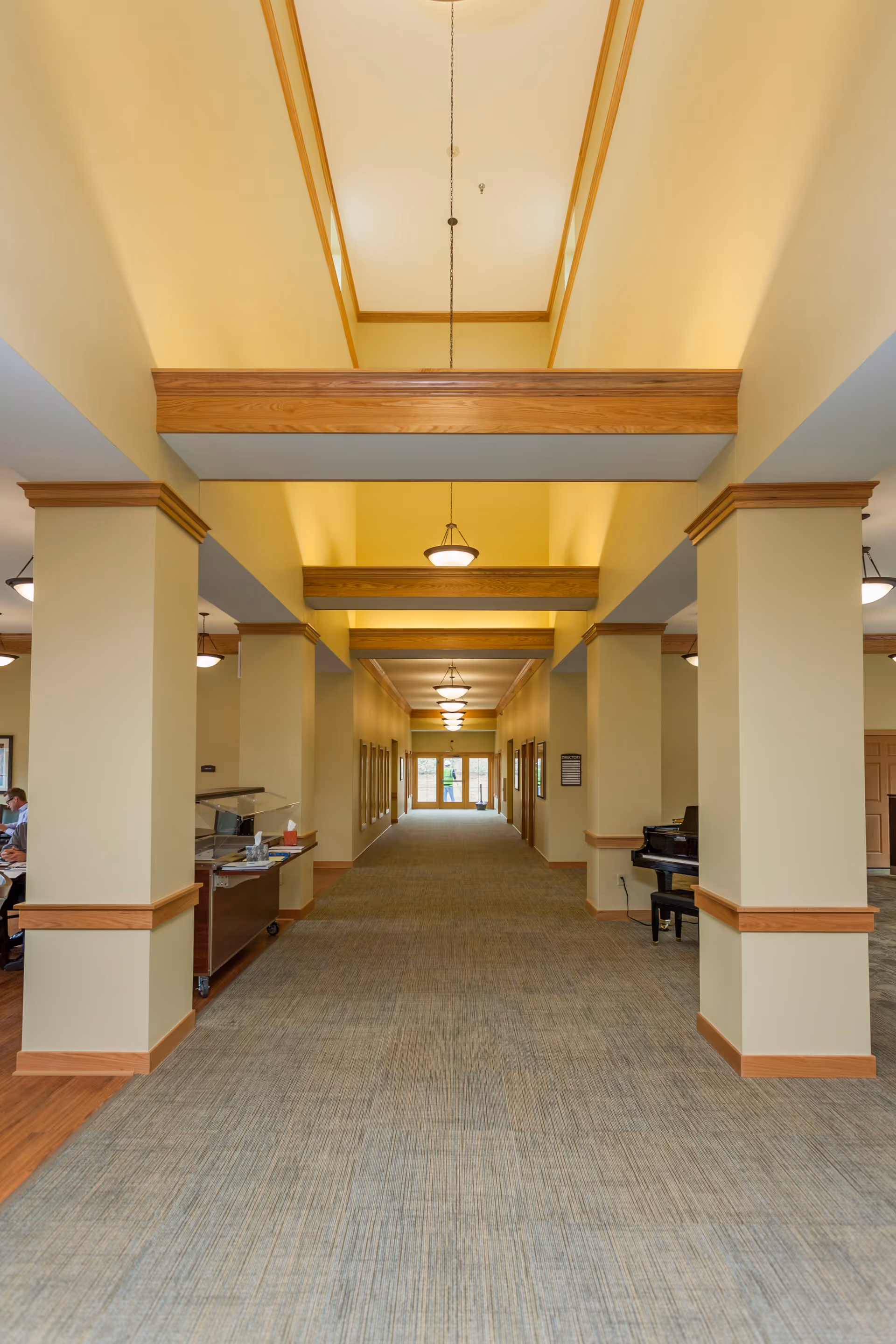 A long, wide hallway in a senior living facility with beige walls and carpeted floor. The hallway features multiple square columns with wooden trim and beams across the ceiling. Several hanging light fixtures illuminate the space. On the right side, there is a black piano and on the left side, a small serving station with a person partially visible sitting at a desk. Large windows are visible at the far end of the hallway.