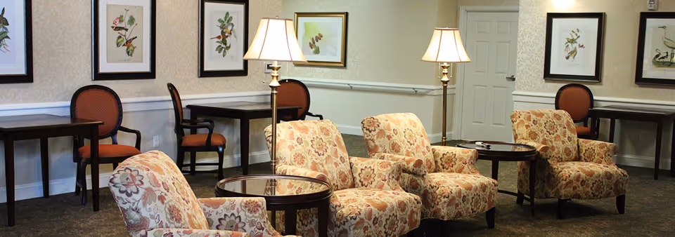 A senior living lounge with floral upholstered armchairs, round side tables, floor lamps, framed botanical prints, and additional chairs and tables along the wall.