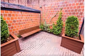 Small outdoor courtyard with brick walls and floor, featuring a wooden bench and three planter boxes with green shrubs.