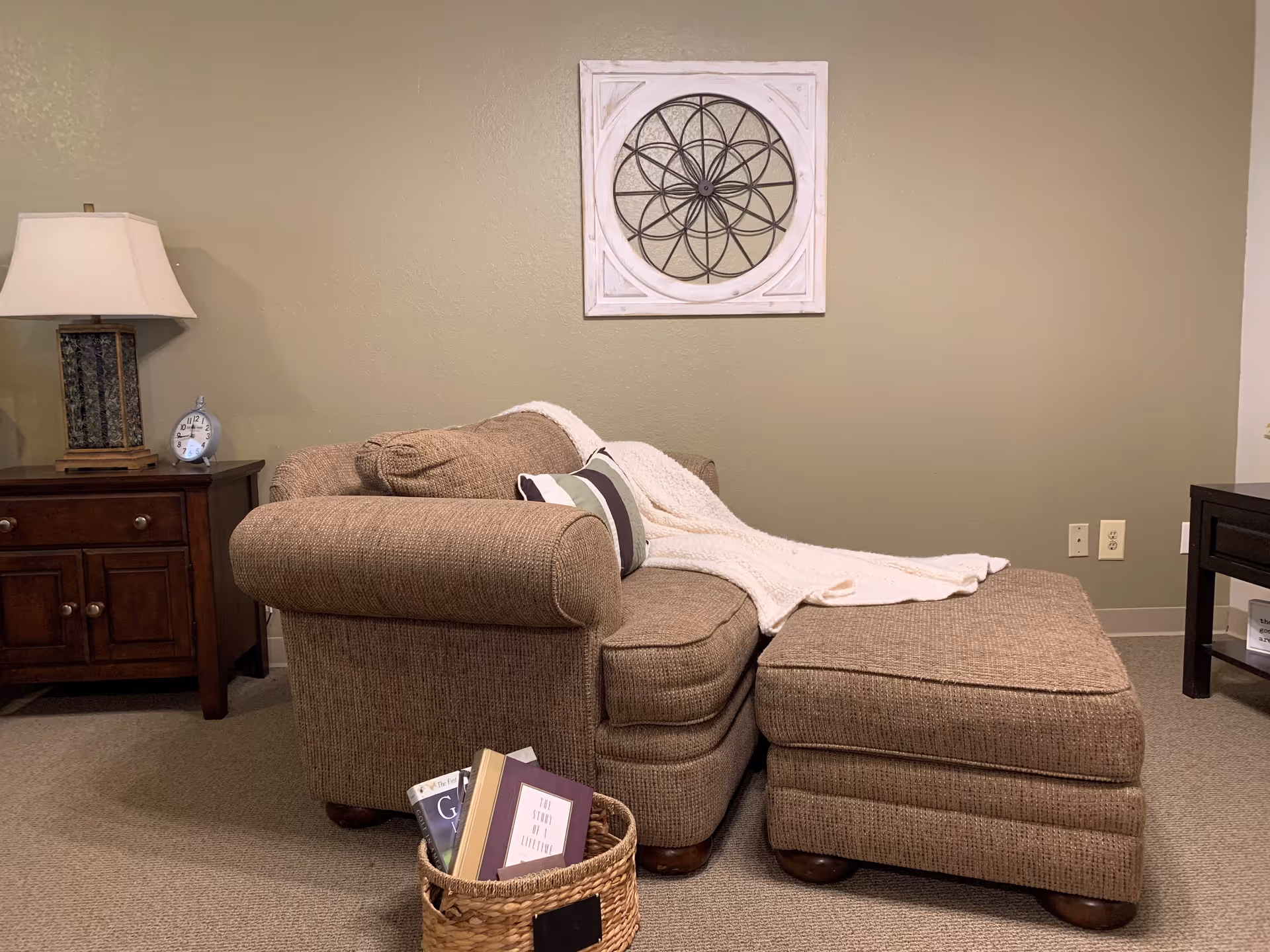 Cozy living room area with a brown upholstered armchair and matching ottoman. A white knitted throw blanket and a striped pillow are placed on the armchair. A wicker basket filled with books sits on the carpeted floor in front of the chair. To the left, there is a wooden side table with a lamp and a small clock. A decorative wall art piece with a circular geometric design hangs on the beige wall behind the chair.