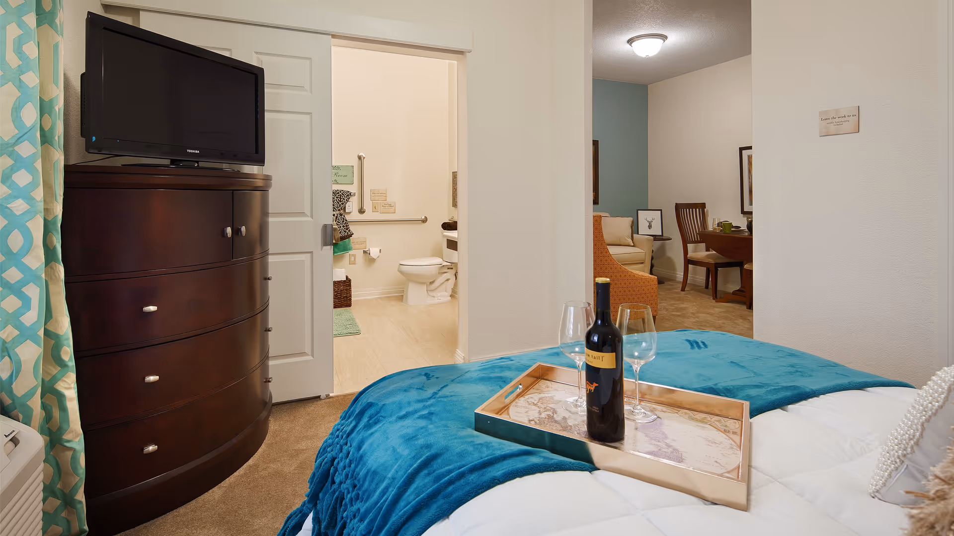A cozy bedroom with a teal blanket on the bed and a wooden tray holding a bottle of wine and two wine glasses. A dark wooden dresser with a TV on top is positioned against the wall. The room opens into a bathroom with a visible toilet and grab bars, and further beyond, a living area with a chair and dining table can be seen.