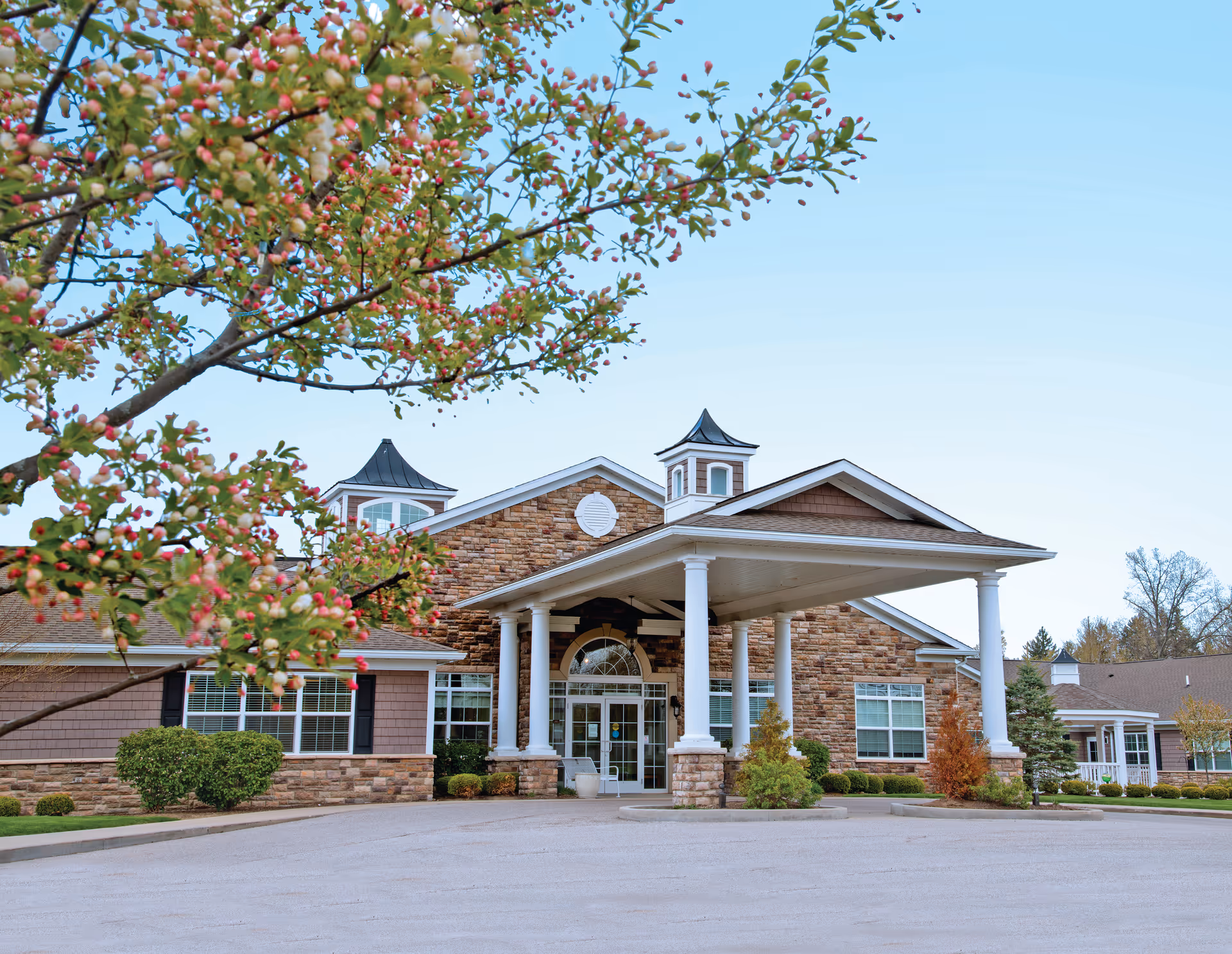 Front exterior view of a senior living facility with stone and siding facade, white columns supporting a covered entrance, and a tree with pink blossoms in the foreground.