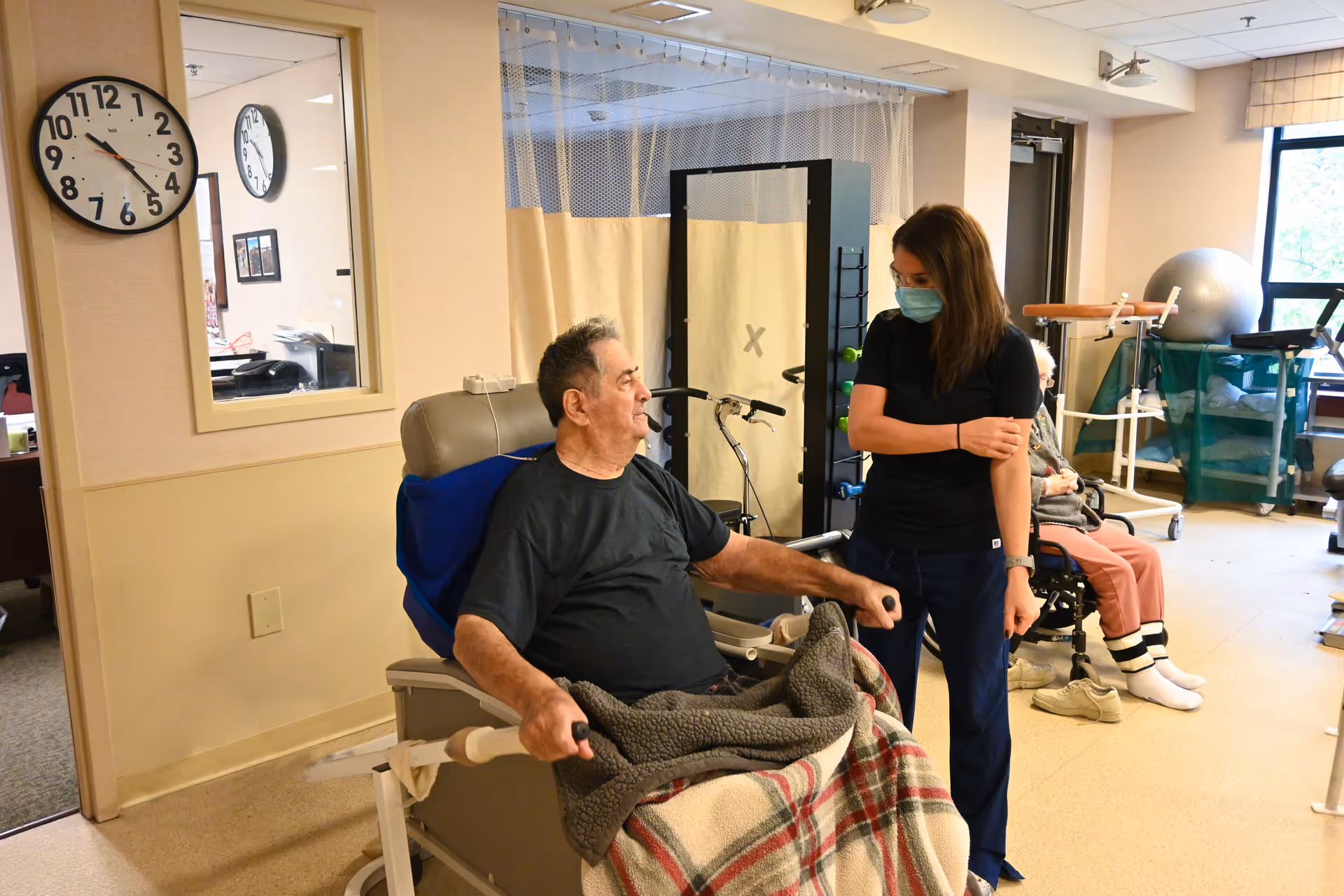 An elderly man sitting in a wheelchair covered with a blanket is interacting with a female caregiver wearing a face mask in a room equipped with exercise and rehabilitation equipment. Another elderly person is seated in the background near a window.