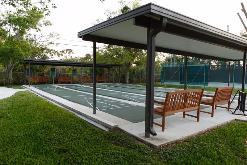 Outdoor shuffleboard courts with covered seating benches and nearby tennis court on a grassy landscaped lot.