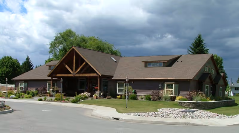 Exterior view of a single-story assisted living facility building with a brown roof and wooden entrance structure, surrounded by green lawns, shrubs, and a stone-bordered garden bed under a cloudy sky.
