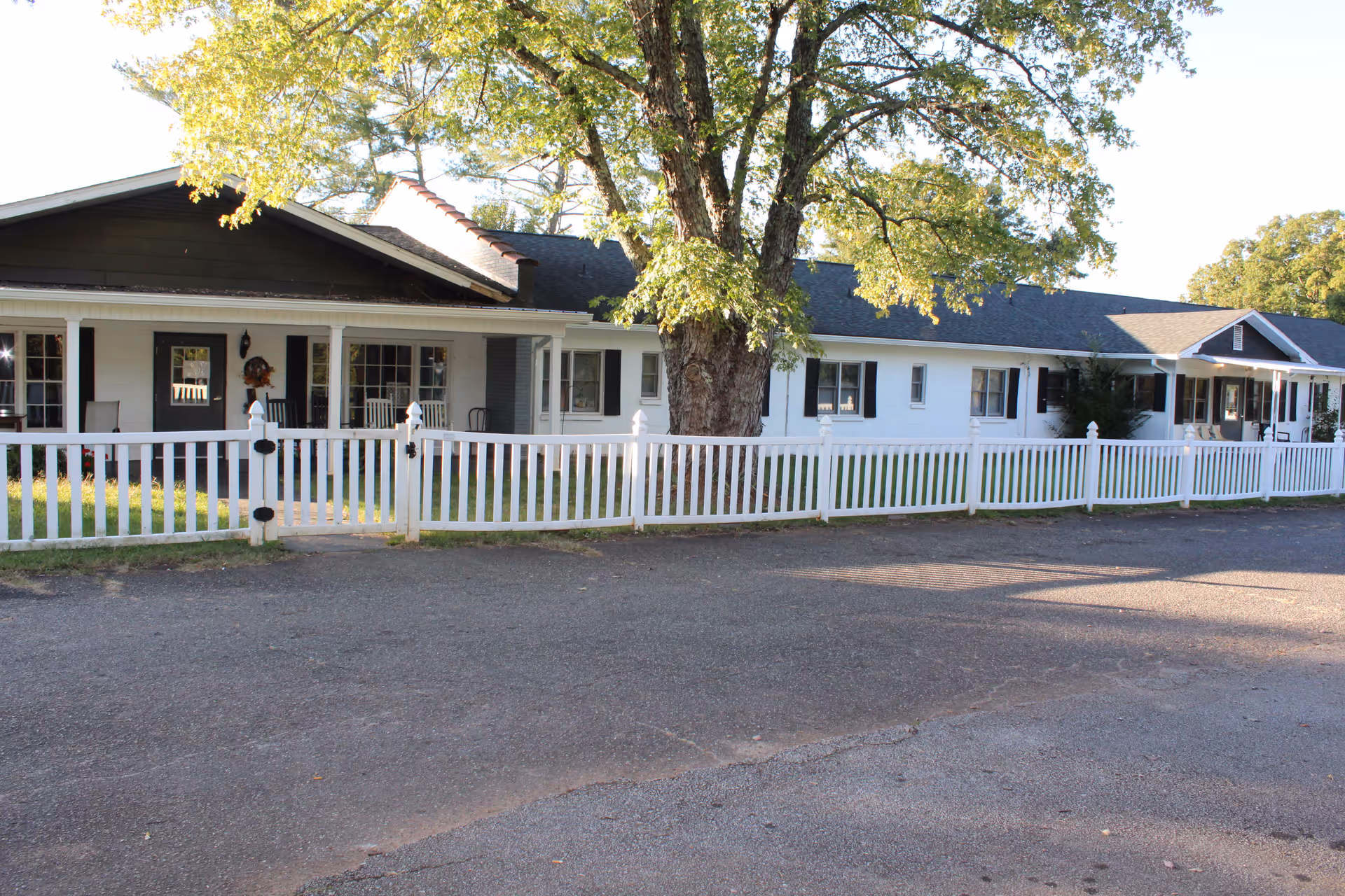 Exterior view of a single-story senior living facility with white walls, black shutters, and a dark roof. The building is surrounded by a white picket fence and has a large tree in front. There is a paved driveway or parking area in the foreground.