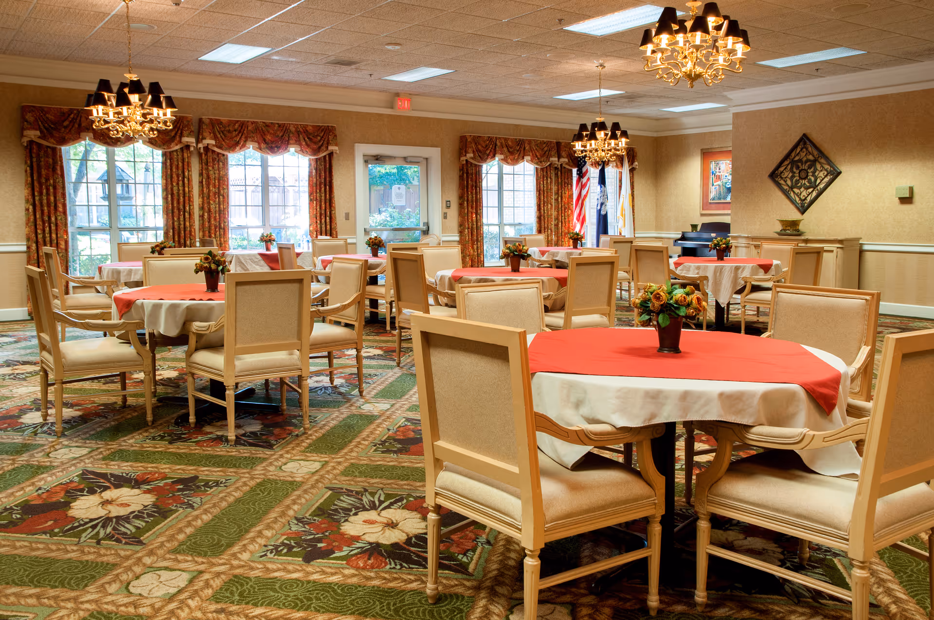 A dining room in a senior living facility with multiple round tables covered with white tablecloths and red overlays. Each table has a small floral centerpiece. The room has beige upholstered chairs, patterned carpet, large windows with floral curtains, and chandeliers hanging from the ceiling.