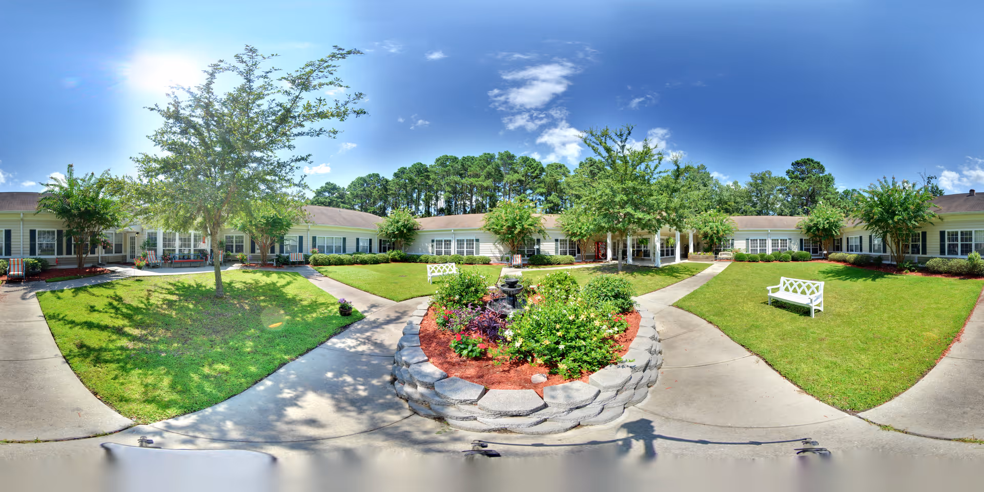 A bright, sunny courtyard at Oaks At Salem Road featuring a circular flower bed with various plants and a small fountain in the center. The courtyard is surrounded by a single-story building with multiple windows and doors. There are several trees and green lawns with white benches placed along the concrete pathways.