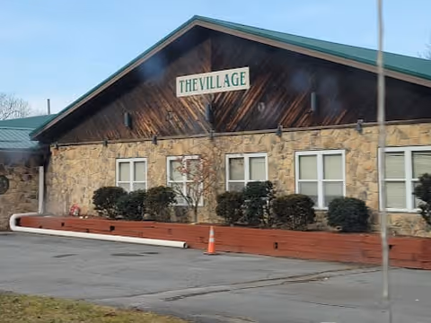 Exterior view of a single-story building with stone facade and wooden upper section. The building has a green roof and a sign that reads 'THE VILLAGE'. There are bushes planted in front of the building and a parking area with an orange traffic cone.