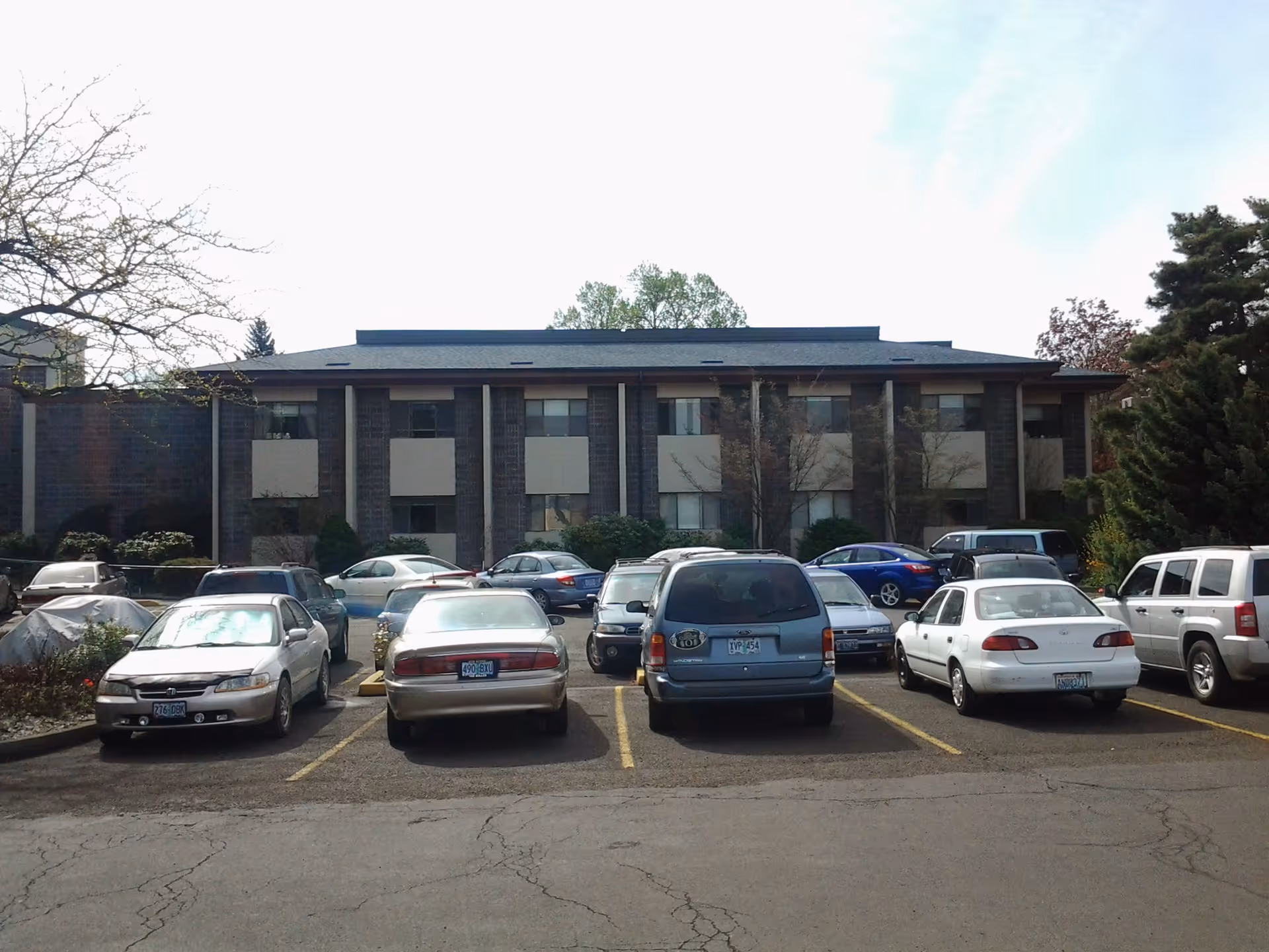 Parking lot with several cars parked in front of a two-story brick building with multiple windows and small balconies, surrounded by trees and shrubs under a partly cloudy sky.