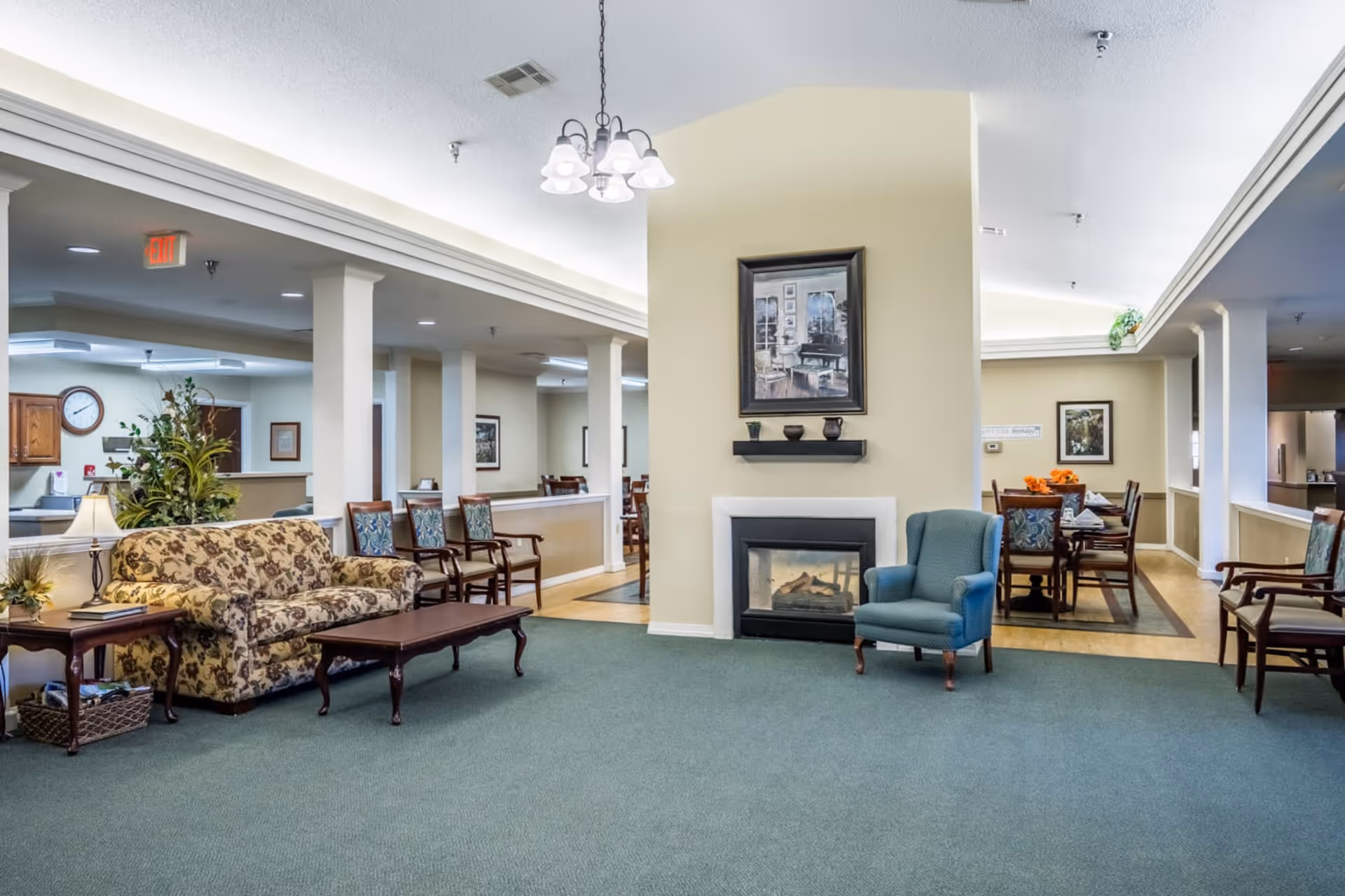 Spacious senior living common area with upholstered seating, a fireplace, and a dining area in the background.