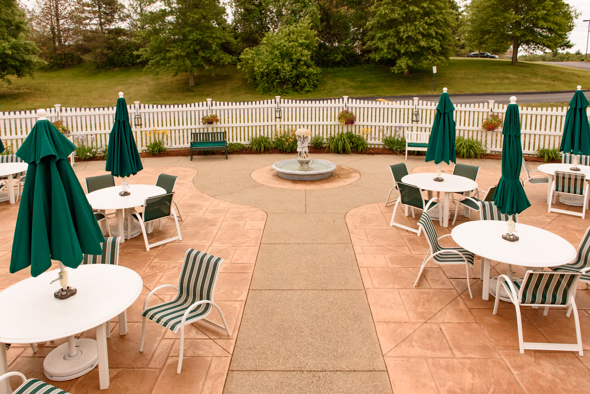 Outdoor patio area with several round white tables and green-striped chairs. Each table has a closed green umbrella. There is a small fountain in the center of the patio, surrounded by a white picket fence and greenery. Trees and a grassy hill are visible in the background.