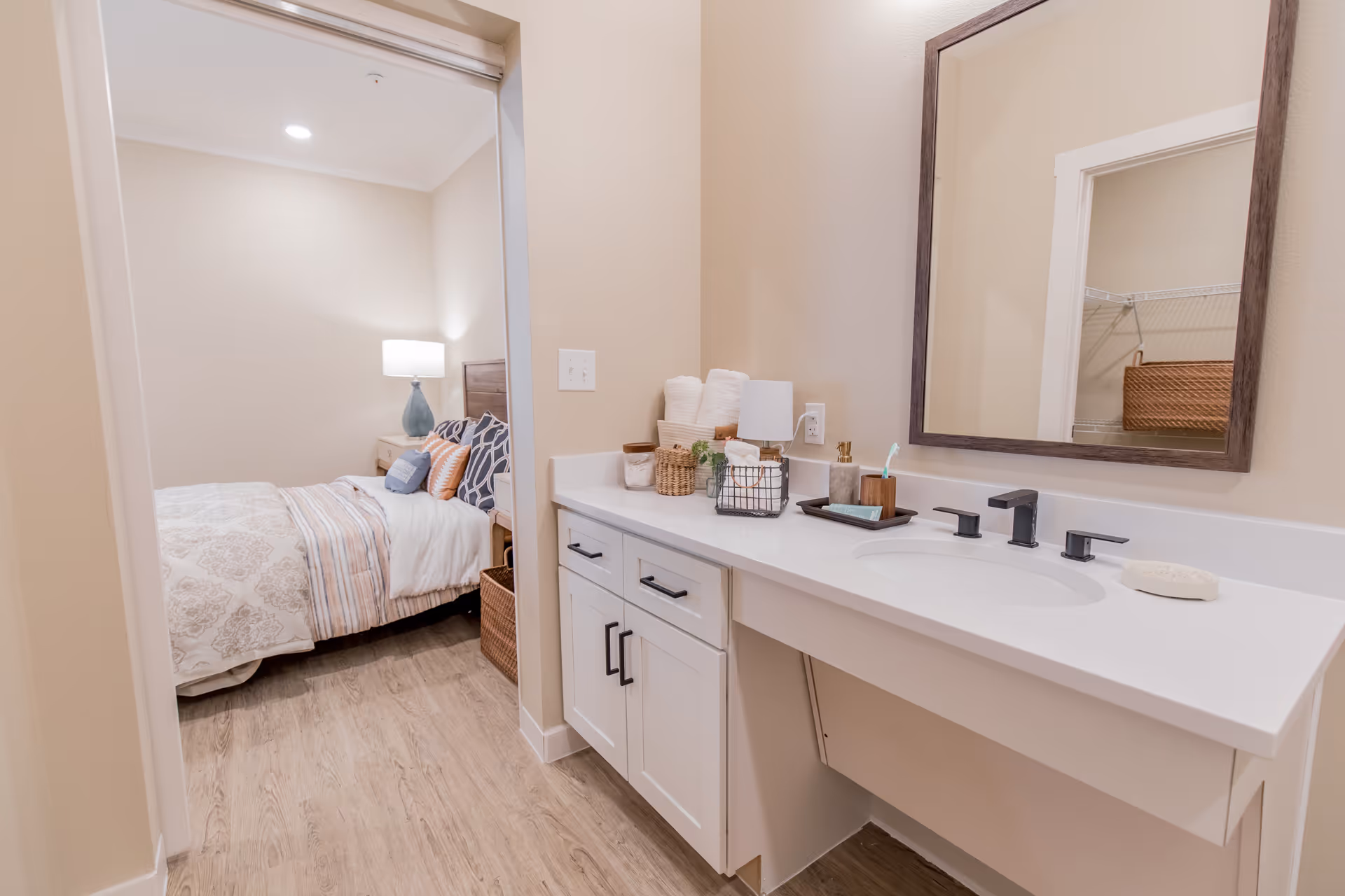 View of a bedroom and adjacent bathroom vanity area in a senior living facility. The bedroom has a bed with patterned bedding, multiple pillows, a nightstand with a lamp, and a wicker basket on the floor. The bathroom vanity features a white countertop with a sink, a large mirror with a dark frame, neatly arranged towels, a soap dish, and various small containers and decor items. The flooring is wood-style, and the walls are painted a light beige color.