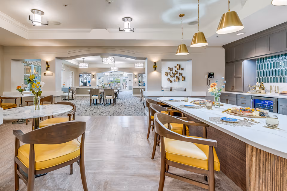 Bright communal dining and kitchen area with an island, bar stools, dining tables, and decorative lighting.