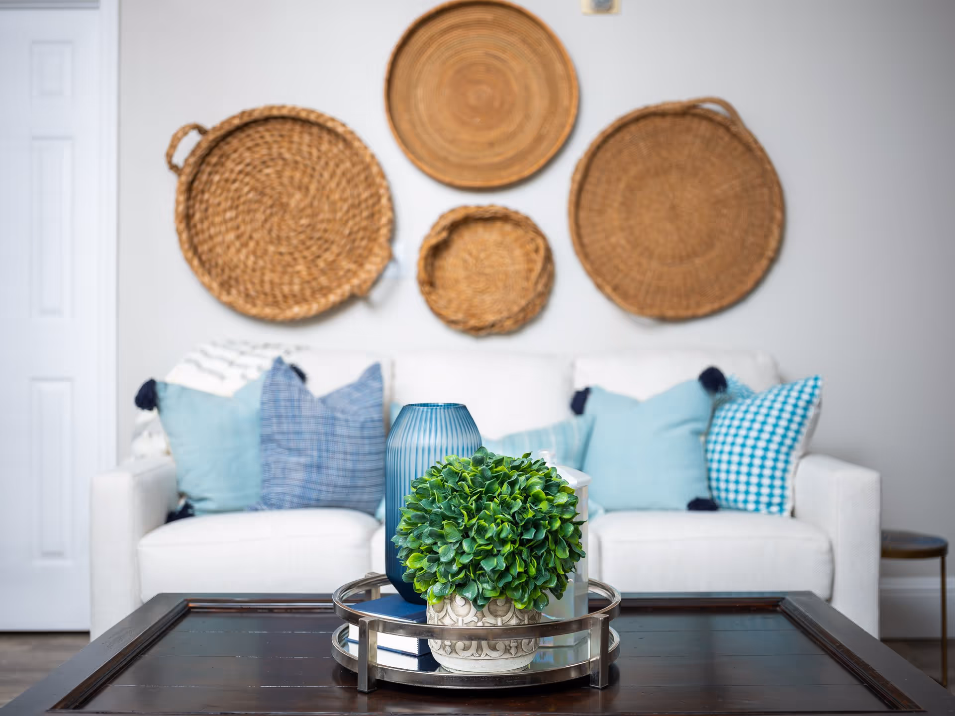 A cozy living room area with a white sofa adorned with various blue and patterned throw pillows. On the wall behind the sofa, there are four decorative woven baskets of different sizes arranged in a cluster. In the foreground, a dark wooden coffee table holds a round metal tray with a small green potted plant, a blue vase, and a white lantern.