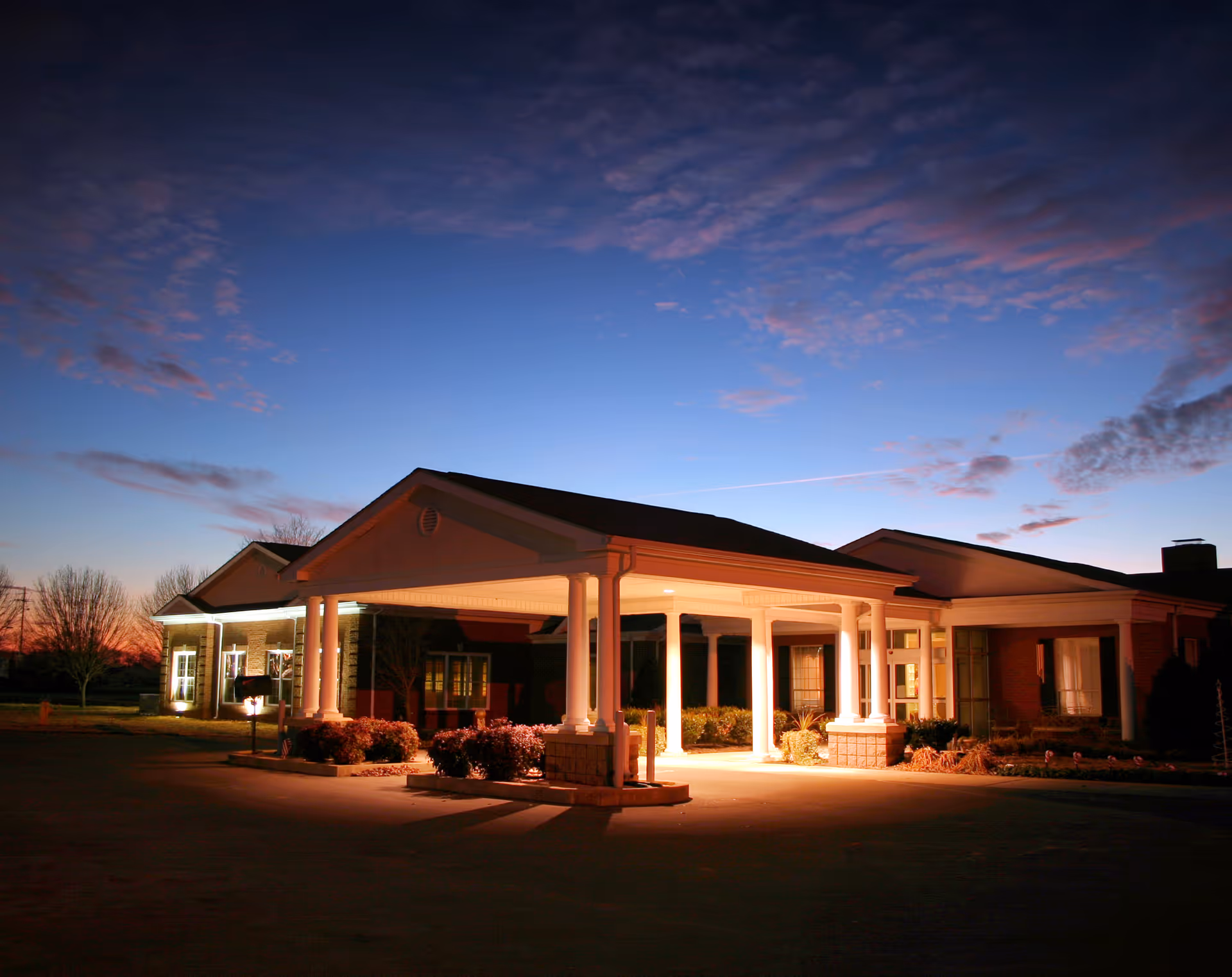 Exterior view of Stones River Manor at dusk with the building entrance illuminated under a covered portico, surrounded by landscaped bushes and trees against a colorful evening sky.