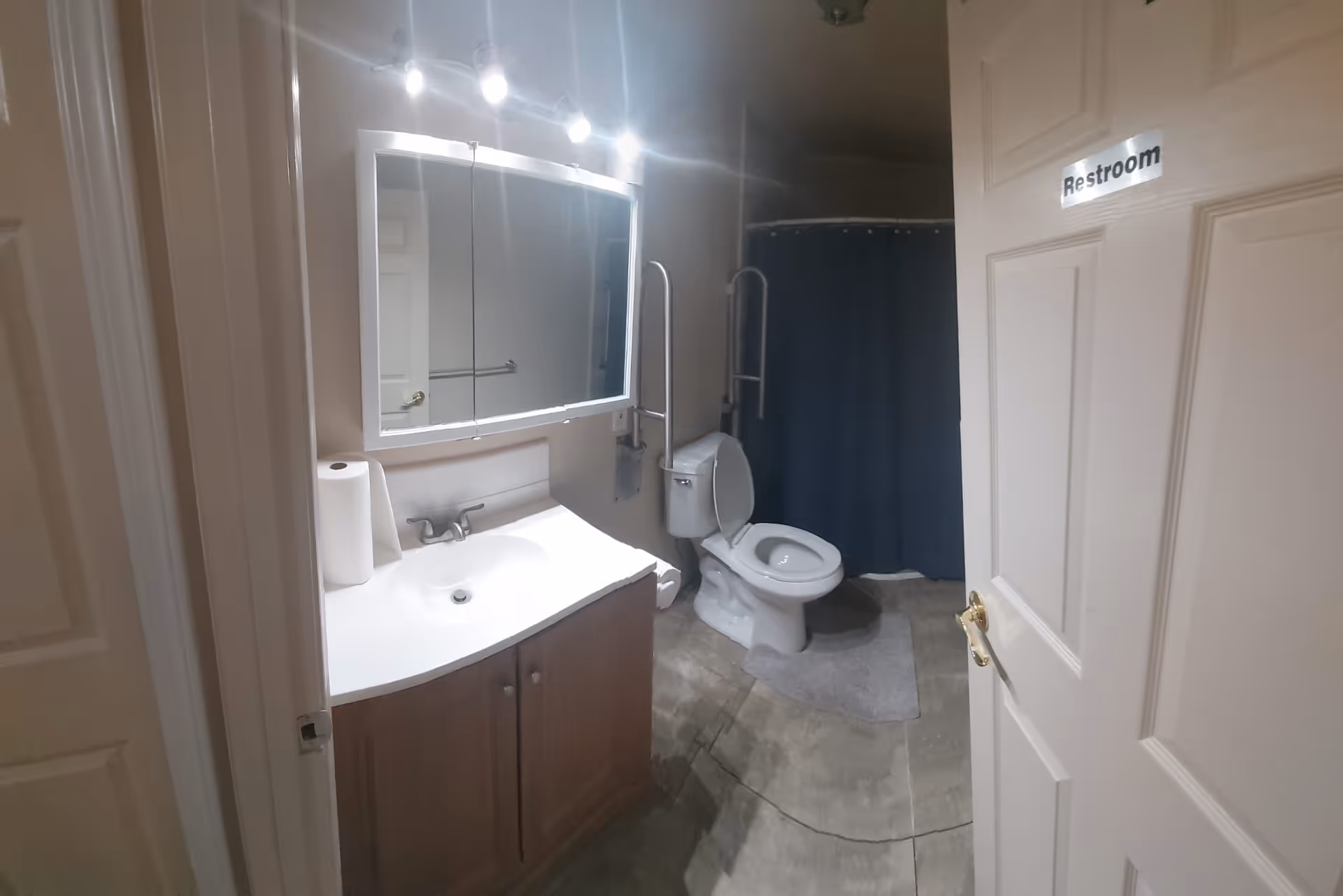 Interior view of a bathroom with a white sink and cabinet, a large mirror with lights above it, a toilet with support bars on both sides, a gray bath mat, and a shower with a dark blue curtain. The door to the bathroom is open and has a sign that reads 'Restroom'.