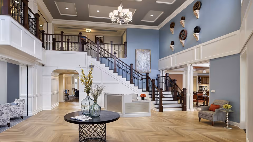 Spacious and elegant interior of an assisted living facility with a large staircase, high ceilings, and decorative wall art. The room features a round table with glass vases and flowers, comfortable seating areas with armchairs, and a chandelier hanging from the ceiling. The walls are painted blue with white wainscoting, and the floor has a light wood herringbone pattern.