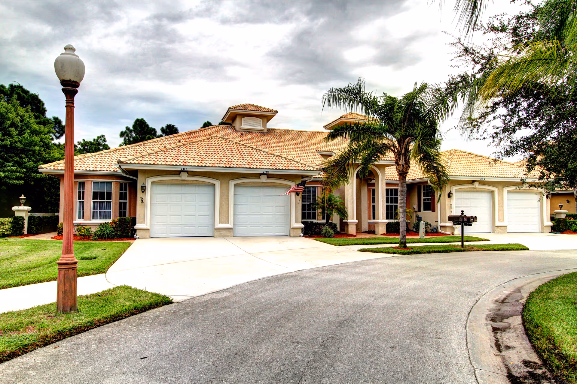 Exterior view of a single-story residential building with a tiled roof, multiple garage doors, palm trees, and a curved driveway under a cloudy sky.
