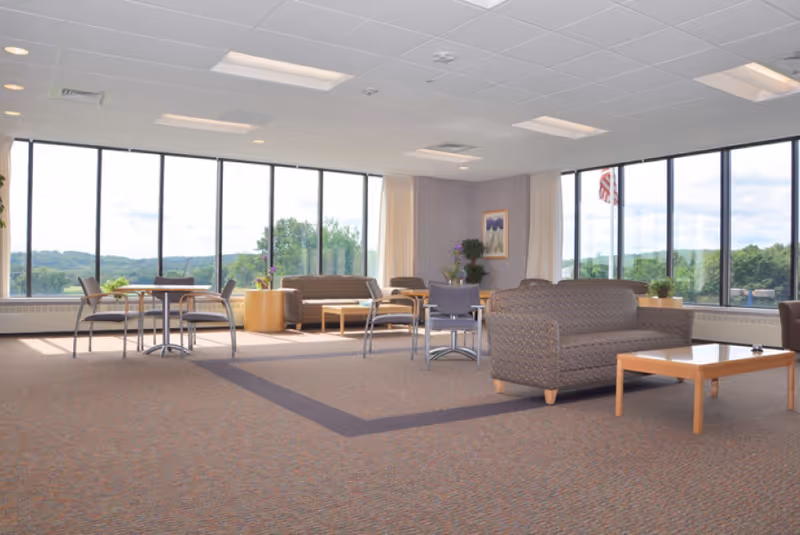 Spacious sunlit common lounge with sofas, chairs, and tables in front of large windows overlooking greenery.