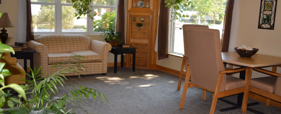 A cozy living room area with a plaid loveseat, a wooden dining table with four upholstered chairs, a small black side table with a lamp, and large windows letting in natural light. There are green plants and a wooden shelving unit in the corner.