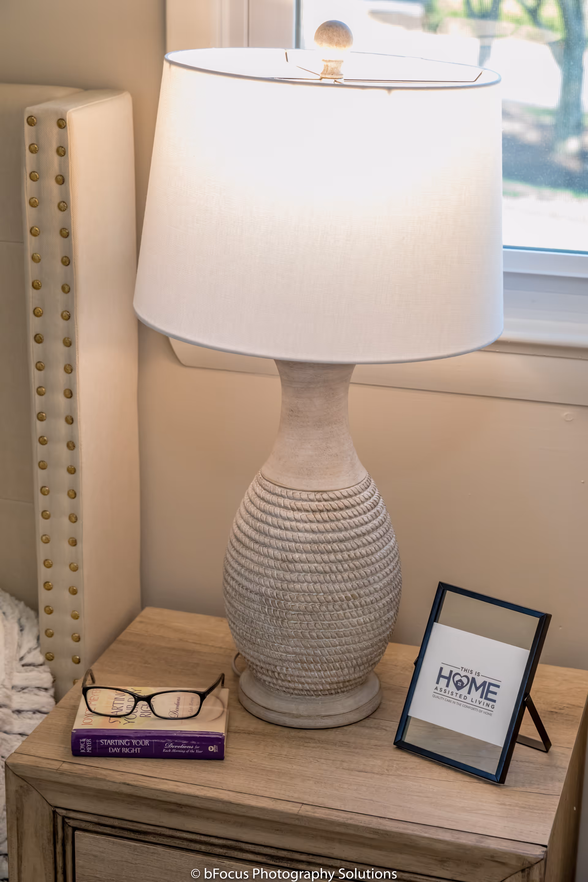 A bedside table with a textured ceramic lamp with a white lampshade, a pair of eyeglasses resting on a book titled 'Starting Your Day Right', and a framed sign that reads 'This is Home Assisted Living'. The table is next to a beige upholstered headboard with brass nailhead trim, and a window is visible in the background.