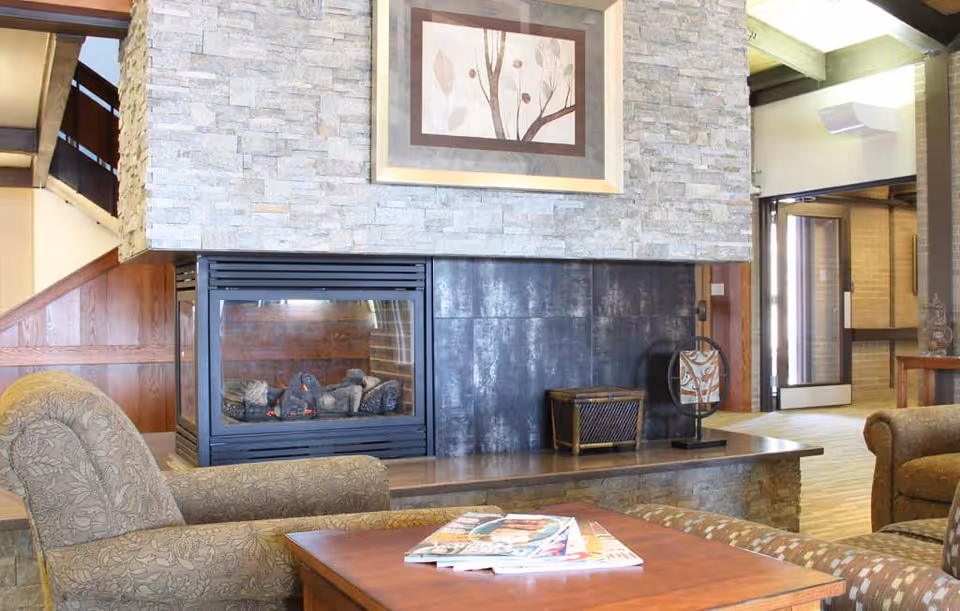 Cozy lounge with upholstered chairs, a coffee table with magazines, and a stone-clad double-sided fireplace.