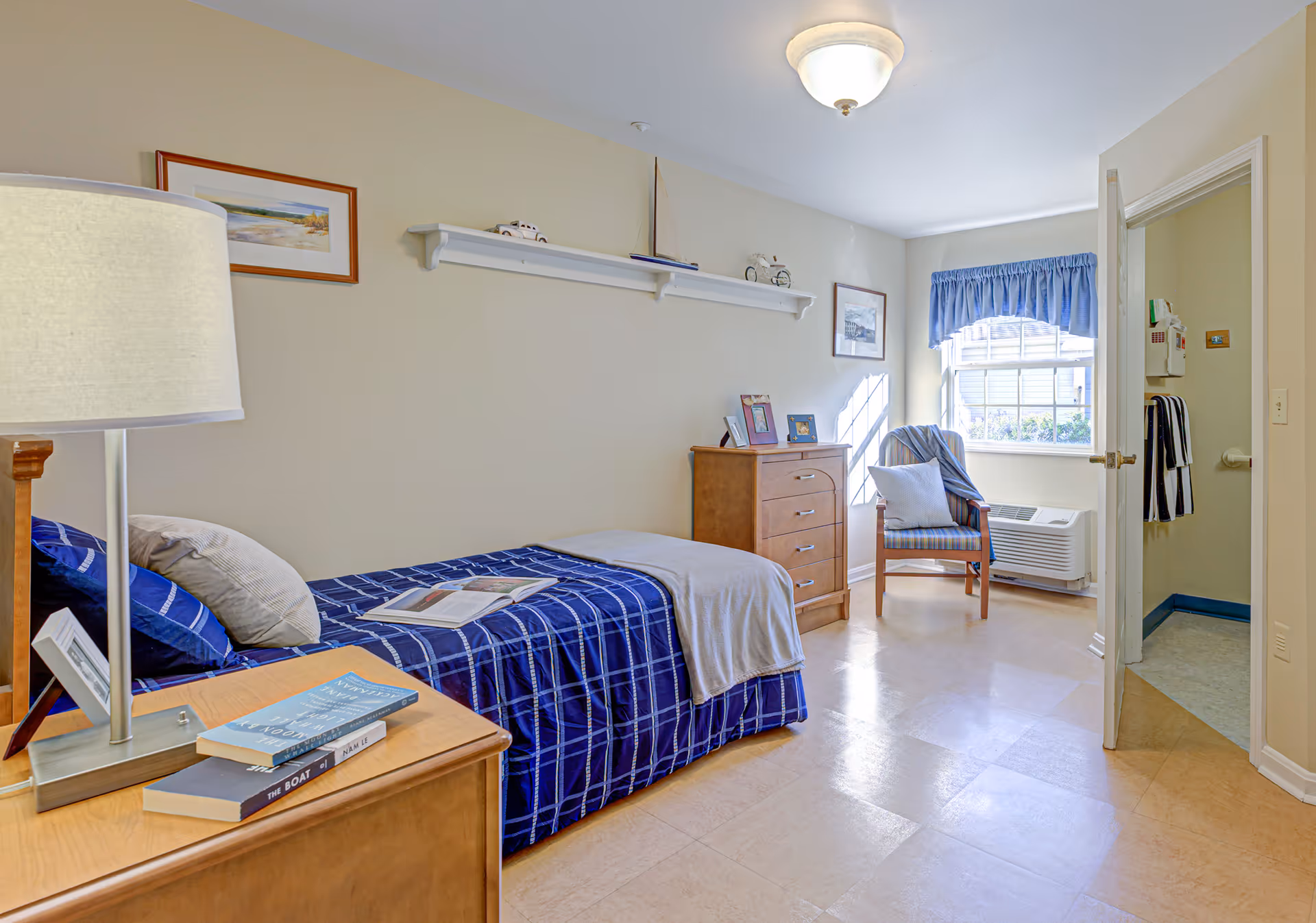 A bright and tidy bedroom in a senior living facility featuring a single bed with blue checkered bedding and a beige throw blanket. Next to the bed is a wooden nightstand with a lamp and books. On the opposite wall, there is a wooden dresser with framed photos and a chair with a cushion near a window with blue curtains. The door to a bathroom is open, showing a towel hanging inside.