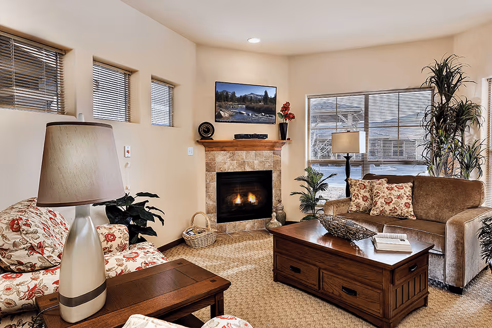 A cozy living room with a floral patterned armchair and a brown sofa adorned with floral cushions. There is a wooden coffee table with decorative items and an open book on it. A fireplace with a tiled surround is lit, and above it hangs a landscape picture. The room has several windows with blinds, a table lamp on a side table, and several green plants adding to the decor.