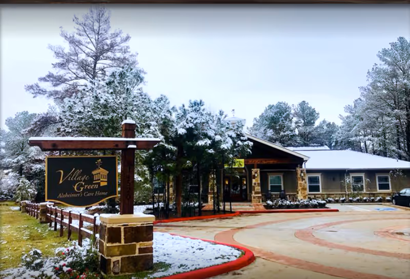 Exterior view of Village Green Memory Care Community Cypress building on a snowy day. The building is surrounded by snow-covered trees and a driveway with red curbs. A wooden sign with the facility name and logo is visible in the foreground.