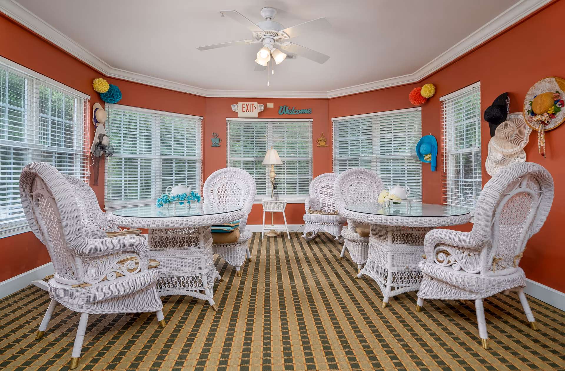 A cozy sitting area with two glass-top wicker tables and six white wicker chairs arranged around them. The room has red walls adorned with various hats and colorful decorations. Large windows with white blinds let in natural light. A ceiling fan with lights hangs above, and a small white side table with a lamp is positioned between the windows. A sign above the windows reads 'Welcome' next to an exit sign.