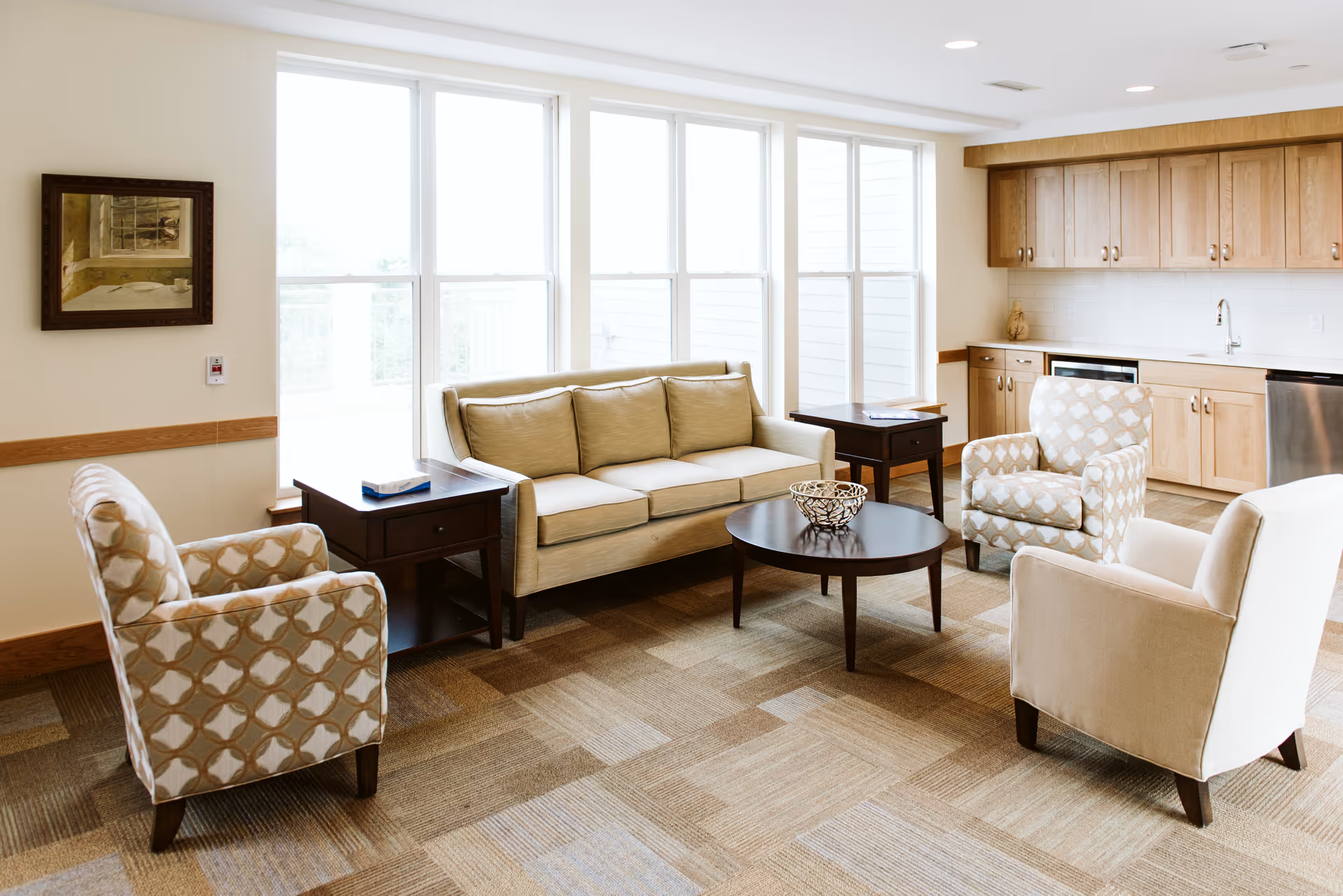 A bright and spacious living room area with large windows letting in natural light. The room features a beige sofa, two patterned armchairs, a plain armchair, a round wooden coffee table with a decorative bowl, and two wooden side tables. In the background, there is a kitchenette with wooden cabinets, a sink, and a dishwasher.