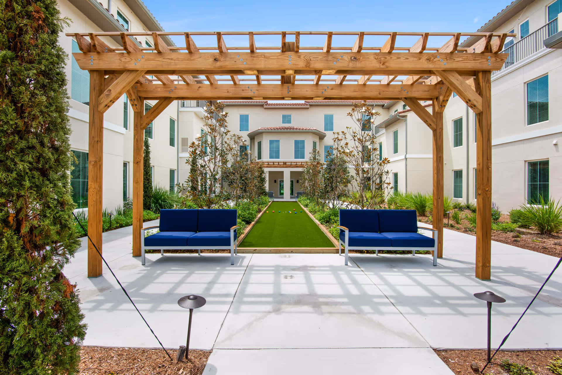 Outdoor courtyard area at Tuscan Gardens Of Delray Beach featuring a wooden pergola with two blue cushioned benches underneath. In the center, there is a bocce ball court surrounded by landscaped plants and trees, with a multi-story building in the background.
